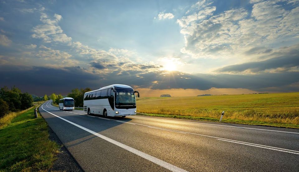 Two white tour buses drive along a highway through a scenic countryside under a partly cloudy sky.